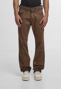 Pantalones de algodón marrones con corte recto, detalles de costura visibles y bolsillos frontales. Llevados con zapatillas blancas y beiges.