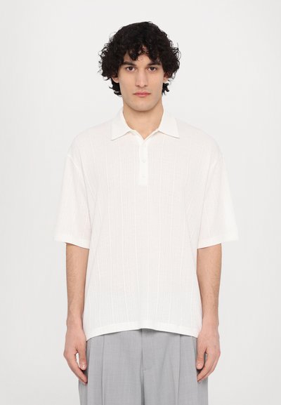 Polo shirt blanc à manches courtes avec un tissu texturé, un col et une patte de boutonnage à trois boutons. Associé à un pantalon plissé gris clair.