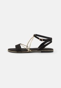 Black suede sandal with gold accents, featuring a flat sole, strappy design, and an adjustable ankle strap. Minimalist and open-toed style.