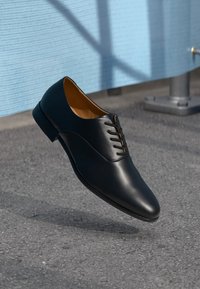 Black leather dress shoe floating above asphalt ground near a light blue wall with metal pipe support.