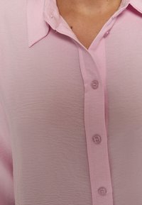 Camicia button-up rosa chiaro realizzata in tessuto testurizzato. Presenta un colletto classico e cinque bottoni trasparenti lungo il patta frontale.