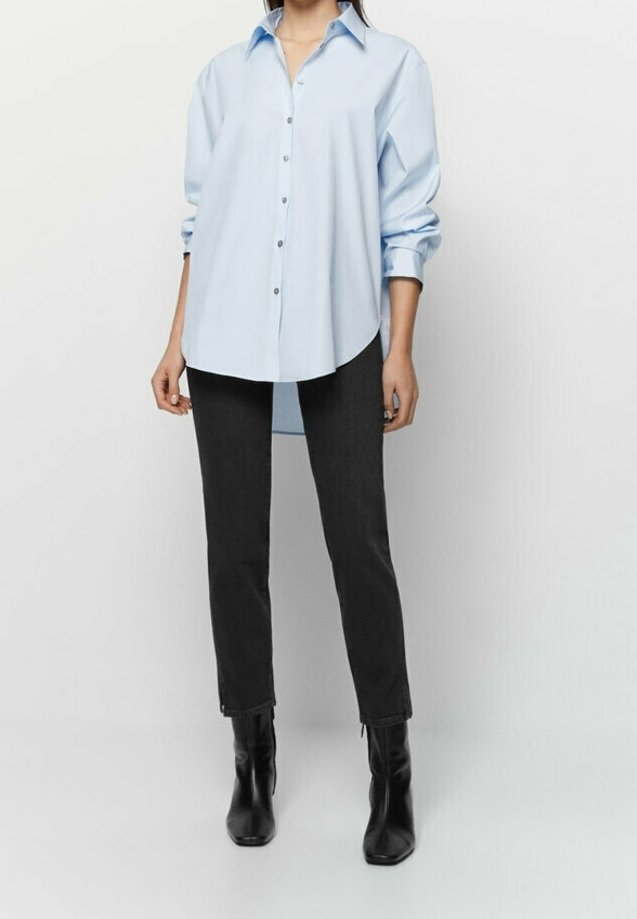 Femme portant une chemise oversize bleu clair boutonnée, un pantalon noir slim et des bottines noires, debout devant un fond blanc uni.