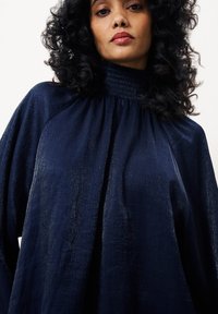 Blouse marine avec un col smocké haut, en tissu fluide et avec un motif texturé subtil. Ce modèle présente des manches larges et une coupe décontractée.