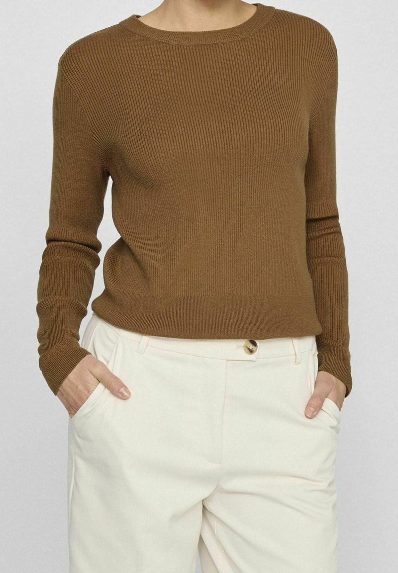 Femme portant un pull côtelé marron ajusté à manches longues et un pantalon taille haute blanc cassé, les mains dans les poches.
