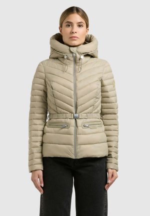 Kobieta w jasnobeżowej pikowanej kurtce puchowej z kapturem, paskiem i zamykanymi na suwak kieszeniami, ubrana w czarne spodnie, stojąca na jednolitym tle.