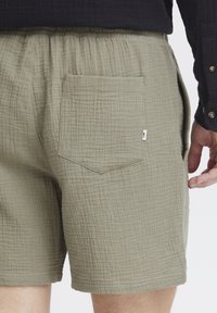 Shorts vert olive fabriqués en tissu texturé et froissé. Dotés d'une taille élastique et d'une poche arrière unique avec un rabat triangulaire.