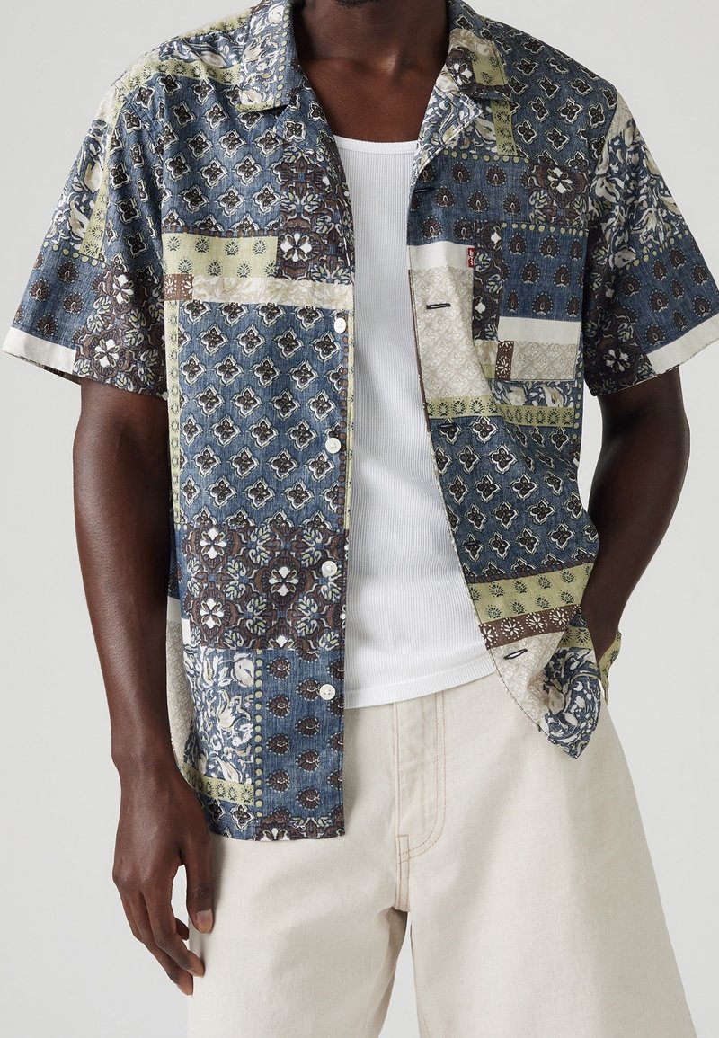 Homme portant une chemise à manches courtes avec boutons, avec un motif patchwork floral bleu, vert et marron mélangé, par-dessus un débardeur blanc côtelé.