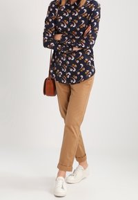 Granatowa koszula na guziki z kwiatowym wzorem, beżowe chinosy i białe sneakersy. Brązowa torba na ramię spoczywa na ramieniu. Rolowane nogawki spodni.