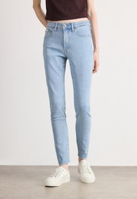 Svetlomodré úzke rifle majú priliehavý strih, päťvreckový dizajn a jemné prané prevedenie. V kombinácii s bielymi teniskami, umiestnené na neutrálnom pozadí.