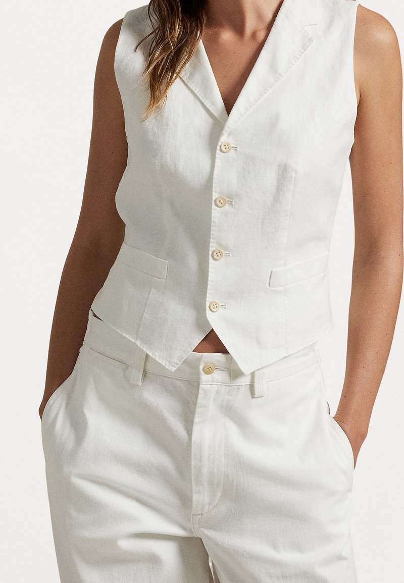 Kvinna klädd i vit ärmlös knäppt väst och matchande vita byxor med händerna i fickorna mot vit bakgrund.