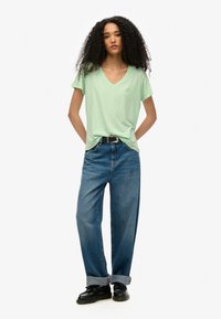 Camiseta verde claro de cuello en v con un logo sutil, combinada con jeans de mezclilla holgados. El conjunto se completa con zapatos negros y un cinturón.