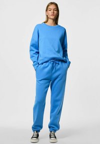 Sudadera de un azul claro y pantalones deportivos de tejido suave, con un corte relajado, puños acanalados y cinturilla ajustable. Zapatillas negras completan el conjunto.