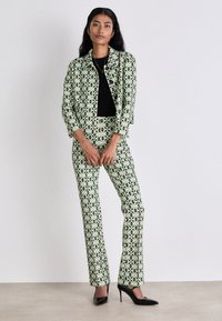 Grüner und schwarzer gemusterter Anzug mit einer kurzen Jacke und eng anliegenden Hosen, die geometrische Sternenmuster aufweisen. Dazu werden schwarze High Heels getragen.