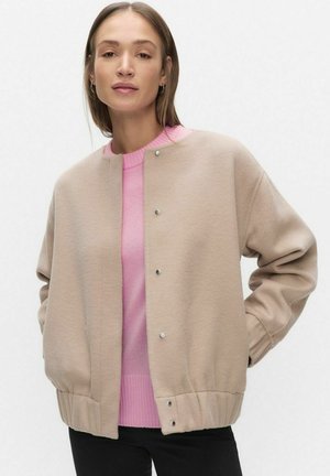 Beige fleecejakke med rund halsudskæring, trykknapper, ribbet kant og oversized ærmer, lagdelt over en lyserød sweater.