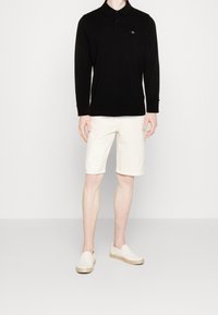 Schwarzes langärmliges Poloshirt mit Kragen und Knopfleiste, kombiniert mit cremefarbenen knielangen Shorts und hellen Slippers.