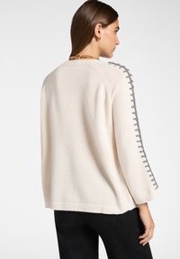 Vrouw met een crèmekleurige trui met grijze stikseldetails langs de mouwen, zwarte broek, gouden ketting en gouden oorringen, zijwaarts gedraaid.