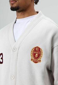 Lichtgrijze sweatvest met een voorzijde met knopen, voorzien van een bordeauxrood geborduurd cijfer "3" en een logopatch met gouden accenten.