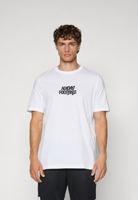 Hvid bomulds t-shirt med korte ærmer, med et sort "Adidas Football" logo i centrum. Rund hals design, afslappet pasform.