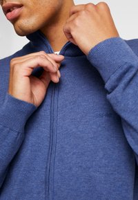 Niebieski sweter zapinany na zamek z wysokim kołnierzem i ściągaczami na mankietach. Tkanina ma fakturowany wygląd, z małym logo na piersi.