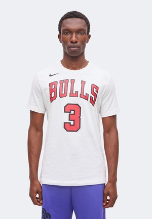 Homme portant un t-shirt blanc avec un texte rouge "BULLS" et le numéro 3, associé à un short violet, debout devant un fond clair uni.