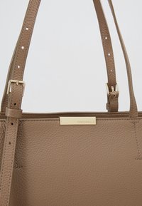 Sac à main en cuir marron avec une finition texturée, doté d'une plaque logo ton or et d'une sangle ajustable avec des accents de boucle.