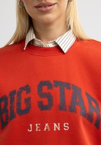 Punane dressipluus koos tekstuuriga sinise trükiga, millel on kiri "BIG STAR JEANS", kantud valge ja musta triibulise kaelusega särgi peale. Sile kangas.