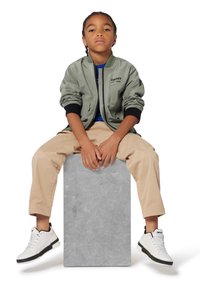 Khaki bomberjack met zwarte accenten, blauwe shirt, beige broek en witte sneakers op een grijze standaard. Eenvoudig ontwerp, casual pasvorm.