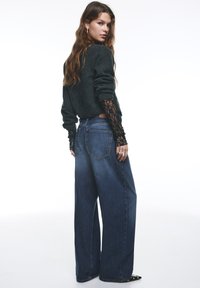 Pull gris foncé avec manches en dentelle et jean bleu à jambes larges avec poches arrière. La tenue a une silhouette décontractée et confortable.