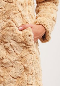 Beige konstläderjacka med en texturerad, plyschyta och en ficka på sidan, vilket ger ett mysigt och varmt intryck.