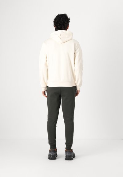 Sweatshirt à capuche en blanc cassé, assorti à un jogging vert foncé. Le tissu semble doux, et le jogging présente une coupe fuselée au niveau de la cheville.