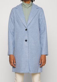 Cappotto in lana celeste con finitura texturizzata, colletto a risvolto, chiusura a un bottone e due tasche frontali. Indossato sopra un dolcevita verde.