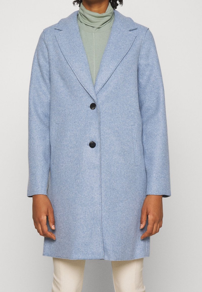 Cappotto in lana celeste con finitura texturizzata, colletto a risvolto, chiusura a un bottone e due tasche frontali. Indossato sopra un dolcevita verde.