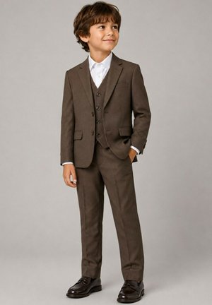 Garçon en costume trois pièces marron avec chemise blanche et chaussures marron foncé, debout une main dans la poche, regardant sur le côté.