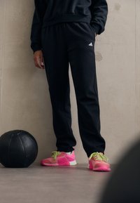 Μαύρο αθλητικό παντελόνι με ελαστική μέση, φορεμένο με ροζ και πράσινα αθλητικά παπούτσια. Ένα μαύρο ιατρικό μπάλα βρίσκεται στο πάτωμα κοντά.