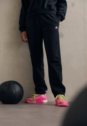 Pantalones deportivos negros con cintura elástica, llevados con zapatillas rosas y verdes. Una pelota medicinal negra yace en el suelo cerca.