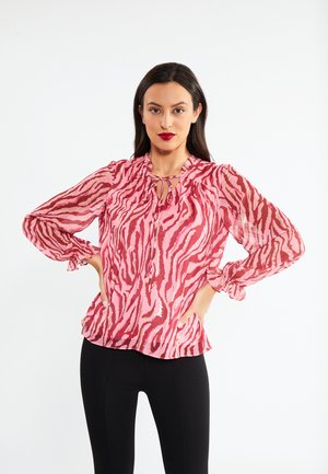 Blusa rosa trasparente con strisce rosse a strisce zebrate, collo rotondo, dettaglio con fiocco e polsini arricciati. Abbinata a pantaloni neri attillati.