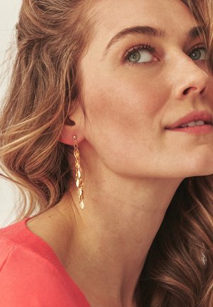 Young woman with wavy light brown hair wears long dangling gold earrings and a coral top, looking slightly upward to the right.
