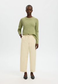 Hellgrüner Strickpullover mit rundem Ausschnitt, kombiniert mit hoch taillierten, beigen, weit geschnittenen Hosen und schwarzen flachen Schuhen. Einfaches und klassisches Design.