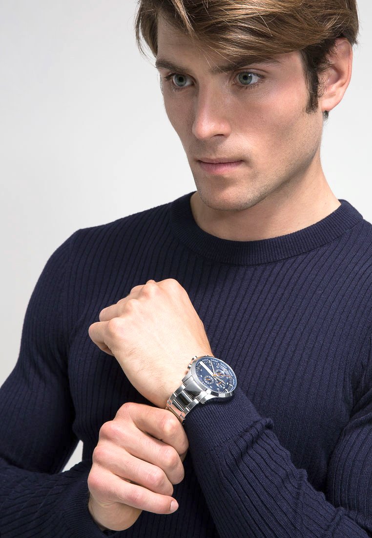 Giovane uomo in maglione blu navy che regola un orologio da polso argentato con quadrante blu, espressione concentrata, contro uno sfondo chiaro e semplice.