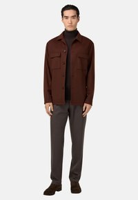 Braunfarbendes Hemd mit Knöpfen und zwei vorderen Taschen, kombiniert mit dunkelgrauen Hosen und braunen Wildlederschuhen. Glatte Textur, lockere Passform.
