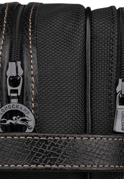 Schwarze, strukturierte Tasche mit zwei Reißverschlüssen, Lederakzenten und kontrastierenden Nähten. Logodetail ist am Reißverschlusszipper sichtbar.