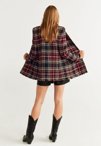 Ruudullinen bleiseri monivärisestä kankaasta, jossa on rispaantuneet reunat. Vaatteena mustien shortsien päällä ja korostettuna mustilla cowboy-kengillä.