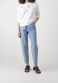 Persona vistiendo una sudadera blanca de manga larga con un pequeño logo en el pecho, jeans de tiro alto azul claro y zapatos de tacón bloque negro de punta abierta.