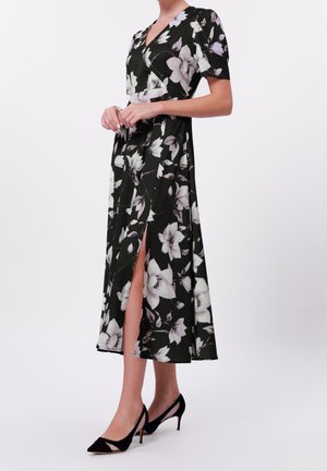 Frau in schwarzem midilangen Kleid mit Blumenmuster, kurzen Ärmeln und seitlichem Schlitz, trägt schwarze spitze Slingback-Absätze, steht vor weißem Hintergrund.
