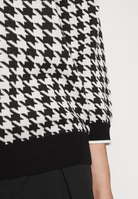 Pull en maille à motif pied-de-poule noir et blanc avec un ourlet et des poignets noirs côtelés ; texture douce ; coupe ajustée ; partie de la manche visible.