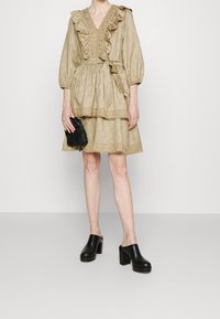 Robe beige avec un col volanté, des manches bouffantes et des dentelles ; associée à des sabots à talons noirs et un petit sac noir.