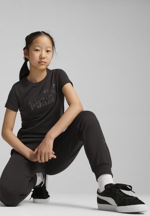 T-shirt noir à manches courtes avec un logo texturé, associé à un pantalon de jogging noir. Des baskets noires et blanches à profil bas complètent la tenue.