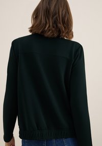 Person med kort brunt hår som bär en svart långärmad tröja med elastisk fåll, sedd bakifrån mot en ljus bakgrund.