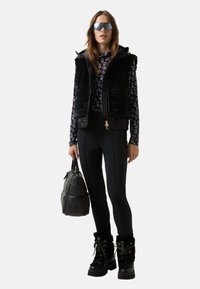 Polstrovaná černá vesta přes vzorovaný černobílý dlouhý rukáv, v kombinaci s černými hladkými legínami a robustními černými botami.