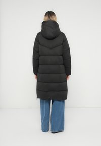 Person set fra baggrunden iført en lang sort hættetrøje og brede blå jeans mod en helt hvid baggrund.